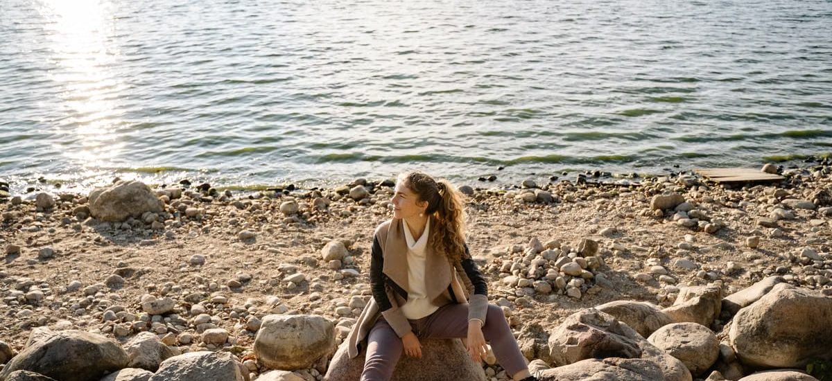 A woman sits on a rocky shoreline, gazing at the serene lake view during a sunny day.