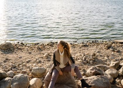 A woman sits on a rocky shoreline, gazing at the serene lake view during a sunny day.